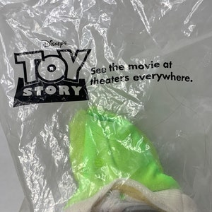 May include: A plastic bag containing a green and white plush toy. The bag has the Disney Pixar Toy Story logo and the text "See the movie at theaters everywhere."