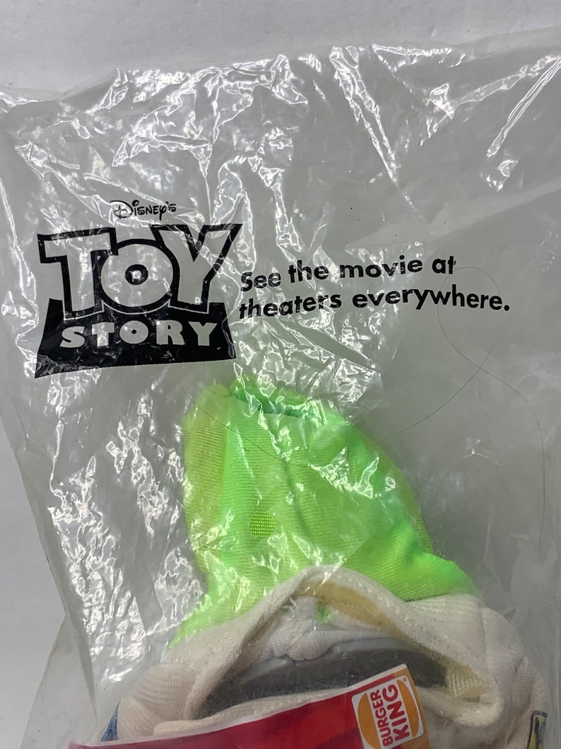May include: A plastic bag containing a green and white plush toy. The bag has the Disney Pixar Toy Story logo and the text "See the movie at theaters everywhere."