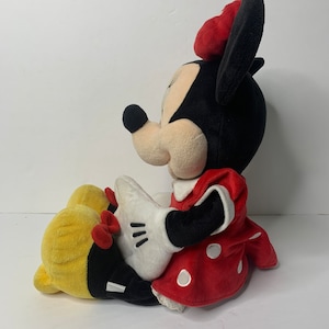 Pu&ograve; includere: Un giocattolo di peluche di Minnie Mouse seduta con le gambe incrociate. Indossa un vestito rosso con pois bianchi e un fiocco rosso. Le sue orecchie sono nere e il suo viso &egrave; bianco con occhi neri e un naso nero.