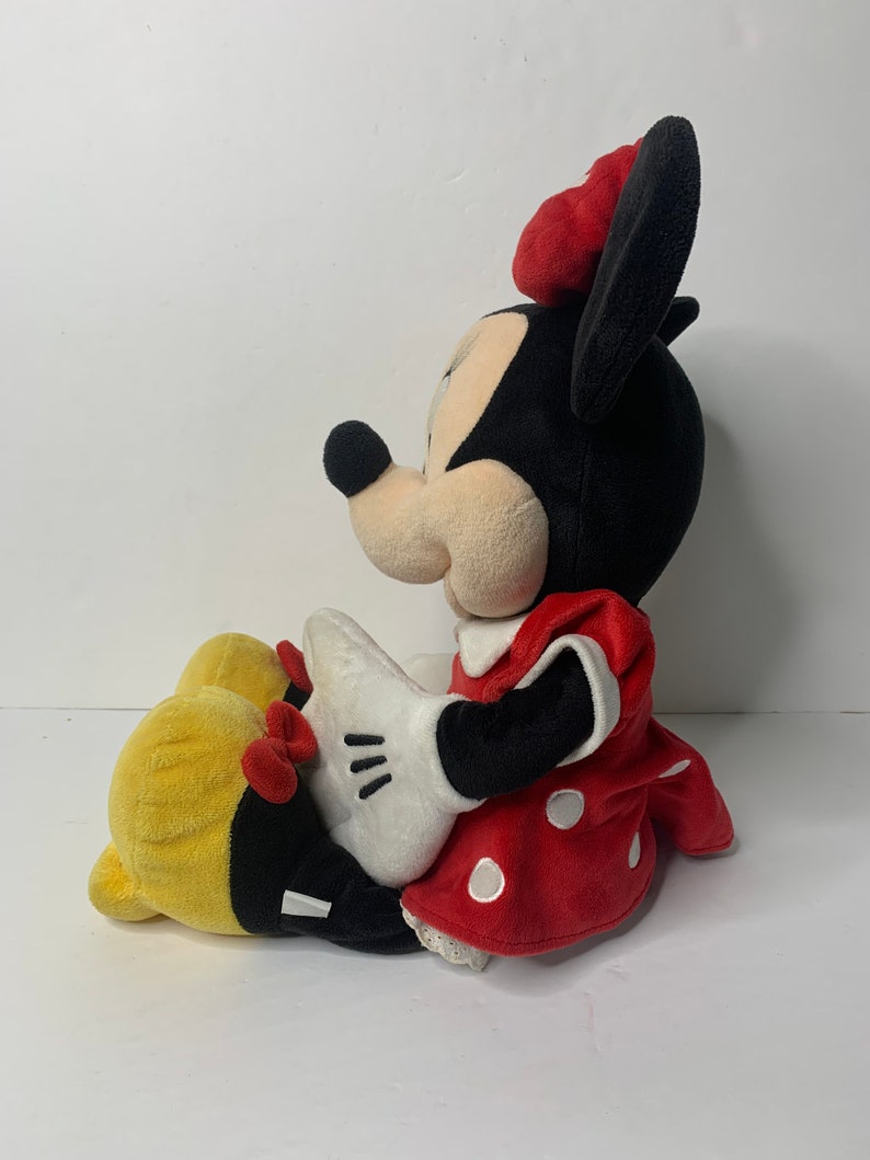 Pu&ograve; includere: Un giocattolo di peluche di Minnie Mouse seduta con le gambe incrociate. Indossa un vestito rosso con pois bianchi e un fiocco rosso. Le sue orecchie sono nere e il suo viso &egrave; bianco con occhi neri e un naso nero.