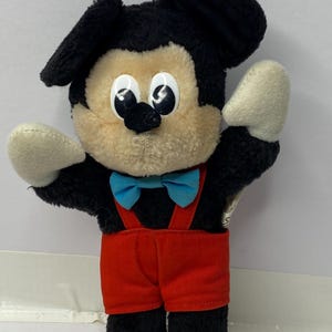 May include: A black and brown plush Mickey Mouse toy wearing red overalls and a blue bow tie. The toy has large, white eyes and a black nose.