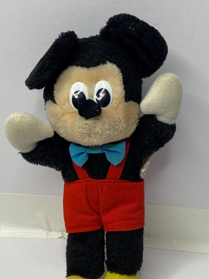 May include: A black and brown plush Mickey Mouse toy wearing red overalls and a blue bow tie. The toy has large, white eyes and a black nose.