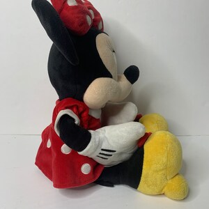Pu&ograve; includere: Un giocattolo di peluche di Minnie Mouse che indossa un vestito rosso con pois bianchi e un fiocco rosso con pois bianchi. Il giocattolo &egrave; seduto con le gambe incrociate e ha uno schema di colori giallo e nero.
