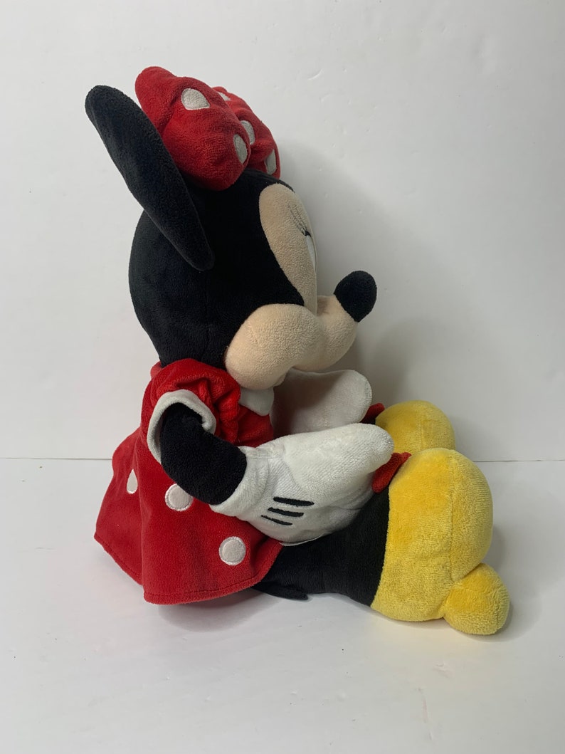 Pu&ograve; includere: Un giocattolo di peluche di Minnie Mouse che indossa un vestito rosso con pois bianchi e un fiocco rosso con pois bianchi. Il giocattolo &egrave; seduto con le gambe incrociate e ha uno schema di colori giallo e nero.