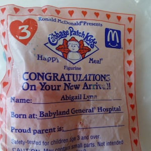 May include: A pink and blue Cabbage Patch Kids Happy Meal toy packaging with a red heart and the number 3. The packaging has a white border with red hearts. The text on the packaging reads "Ronald McDonald Presents Cabbage Patch Kids Happy Meal Figurine Congratulations On Your New Arrival! Name: Abigail Lyna Born at: Babyland General Hospital Proud parent is: Safety-tested for children age 3 and over. CAUTION May contain small parts. Not intended"
