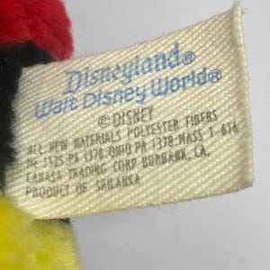 May include: A close-up of a tag from a Disney product. The tag is off-white with blue text that reads "Disneyland Walt Disney World" and "&copy; DISNEY". The tag also includes material and manufacturing information.