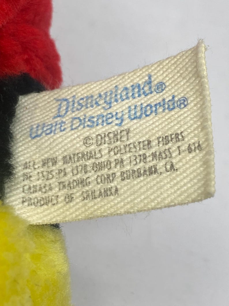 May include: A close-up of a tag from a Disney product. The tag is off-white with blue text that reads "Disneyland Walt Disney World" and "&copy; DISNEY". The tag also includes material and manufacturing information.