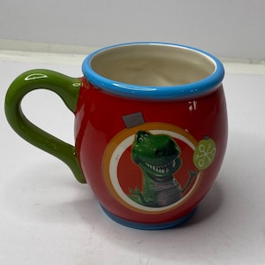 Puede incluir: Una taza de cer&aacute;mica roja con un asa verde y un borde azul. La taza presenta una imagen de dibujos animados de un personaje de dinosaurio verde. Un peque&ntilde;o adorno redondo verde est&aacute; en el lado derecho de la imagen del dinosaurio.