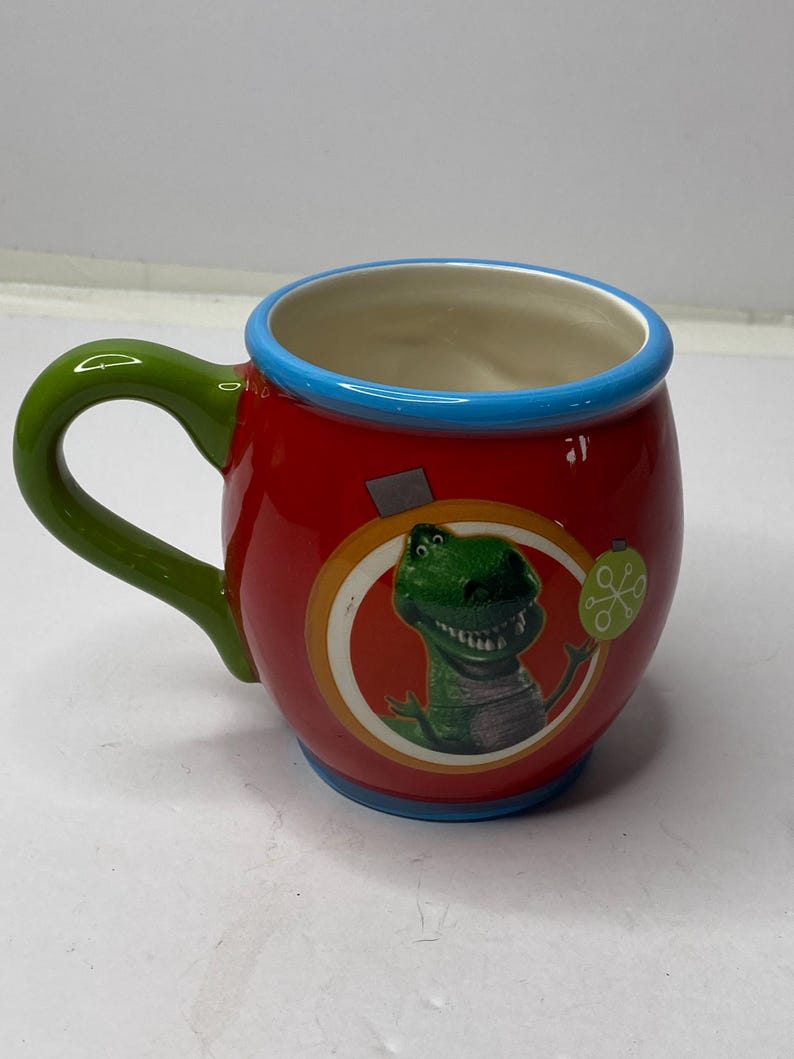 Puede incluir: Una taza de cer&aacute;mica roja con un asa verde y un borde azul. La taza presenta una imagen de dibujos animados de un personaje de dinosaurio verde. Un peque&ntilde;o adorno redondo verde est&aacute; en el lado derecho de la imagen del dinosaurio.