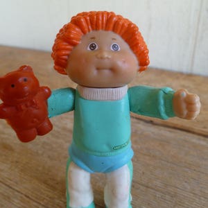 Puede incluir: Una muñeca de pelo rojo con una camisa azul y pantalones blancos sostiene un oso de peluche marrón. La muñeca tiene una cara amigable con ojos azules y una gran sonrisa.