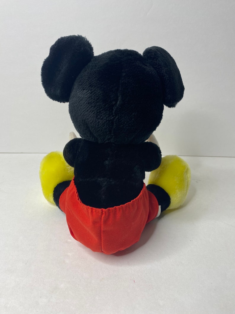 May include: A black and red plush Mickey Mouse toy sitting down with yellow shoes.