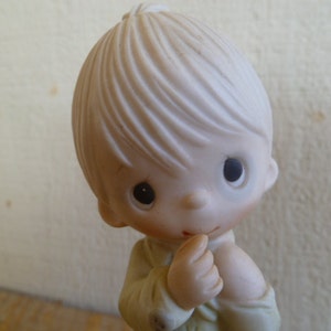 Op de afbeelding: Een porseleinen beeldje van een jonge jongen met blond haar, die een wit shirt draagt en met een bedachtzame uitdrukking naar beneden kijkt.