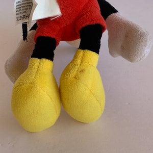 Op de afbeelding: Een Mickey Mouse pluche speelgoed met rode shorts, zwarte benen en gele voeten. De zwarte staart en witte handschoenen van het speelgoed zijn ook zichtbaar. Een label met tekst is bevestigd.