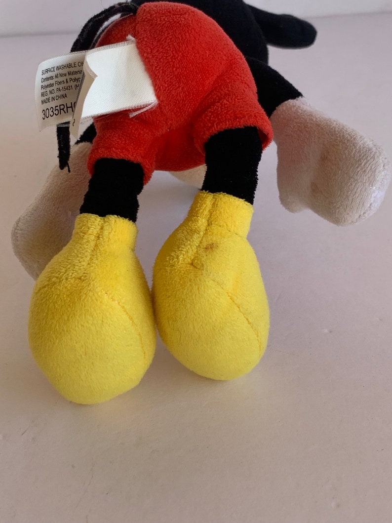 Op de afbeelding: Een Mickey Mouse pluche speelgoed met rode shorts, zwarte benen en gele voeten. De zwarte staart en witte handschoenen van het speelgoed zijn ook zichtbaar. Een label met tekst is bevestigd.