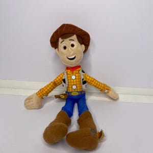 May include: A plush toy of a cowboy character, featuring a brown hat, yellow plaid shirt, blue pants, and brown boots. The toy has a smiling face and is wearing a red neckerchief. The toy is a character from a popular animated movie.