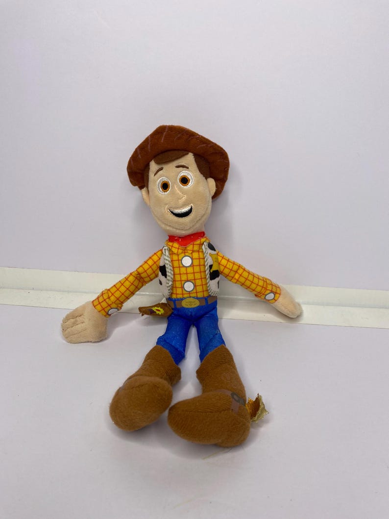 May include: A plush toy of a cowboy character, featuring a brown hat, yellow plaid shirt, blue pants, and brown boots. The toy has a smiling face and is wearing a red neckerchief. The toy is a character from a popular animated movie.