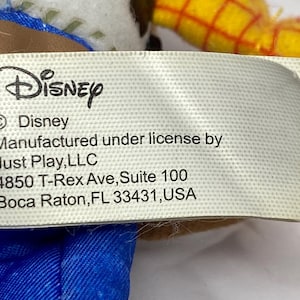 May include: A close-up of a tag with the Disney logo and text. The tag reads "Manufactured under license by Just Play, LLC, 4850 T-Rex Ave, Suite 100, Boca Raton, FL 33431, USA."