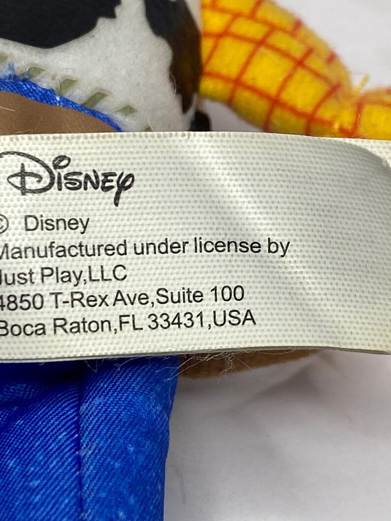 May include: A close-up of a tag with the Disney logo and text. The tag reads "Manufactured under license by Just Play, LLC, 4850 T-Rex Ave, Suite 100, Boca Raton, FL 33431, USA."