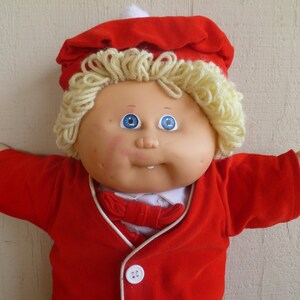 May include: A Cabbage Patch Kid doll wearing a red velvet jacket with white trim and a white bow tie. The doll has blonde yarn hair and blue eyes.
