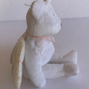 May include: White plush teddy bear with iridescent wings and a pink ribbon around its neck. The bear is sitting with its legs crossed.