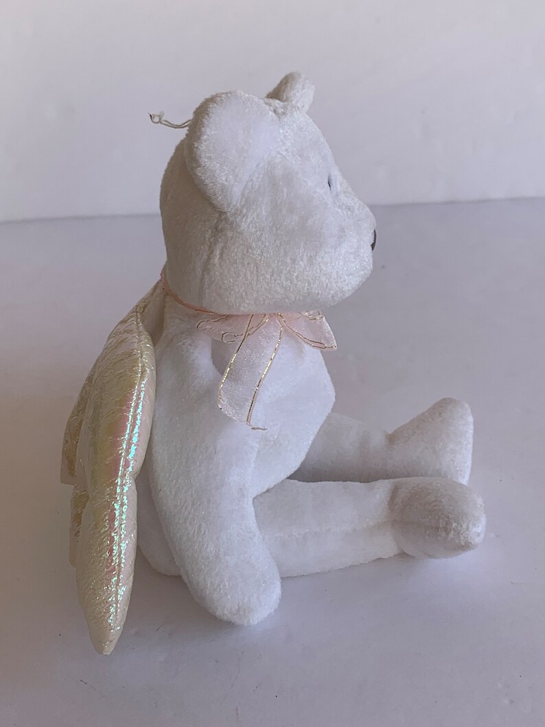 May include: White plush teddy bear with iridescent wings and a pink ribbon around its neck. The bear is sitting with its legs crossed.