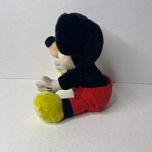 May include: A plush Mickey Mouse toy sitting with his legs crossed. He is wearing red pants with black suspenders and yellow shoes.