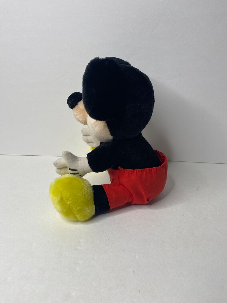 May include: A plush Mickey Mouse toy sitting with his legs crossed. He is wearing red pants with black suspenders and yellow shoes.
