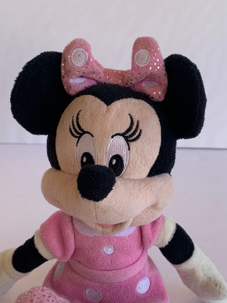 May include: A pink and black plush toy of Minnie Mouse wearing a pink bow with white polka dots and a pink dress with white polka dots.