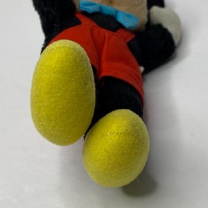 May include: A plush Mickey Mouse toy with black fur, red overalls, and yellow shoes. The toy is lying on its back with its head turned to the side.