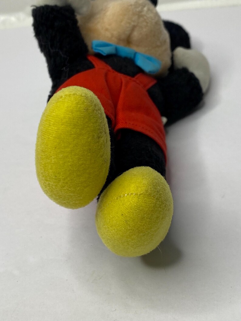 May include: A plush Mickey Mouse toy with black fur, red overalls, and yellow shoes. The toy is lying on its back with its head turned to the side.