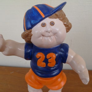 May include: A ceramic figurine of a child wearing a blue and orange baseball cap, a blue jersey with the number 23, and orange shorts.