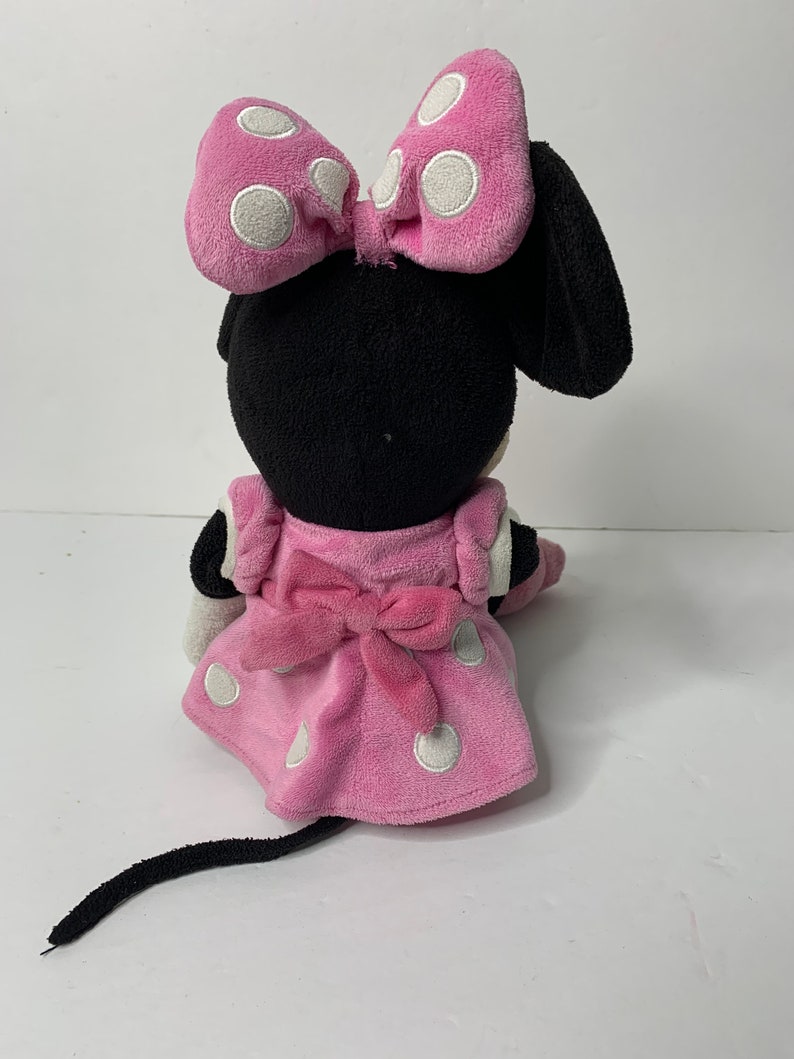 May include: A pink and black plush toy of Minnie Mouse. She is wearing a pink dress with white polka dots and a pink bow. She has a black tail and a black bow on her head.