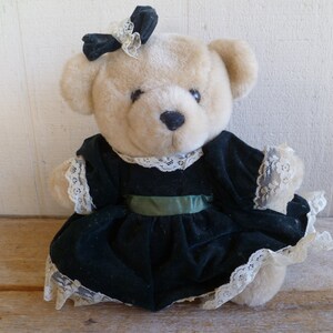 May include: A plush teddy bear wearing a green velvet dress with white lace trim and a black bow. The bear has a light brown fur and black eyes.
