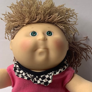 Könnte beinhalten: Eine Cabbage Patch Kid Puppe mit blonden Garnhaaren, grünen Augen und einem pink-schwarz karierten Outfit.