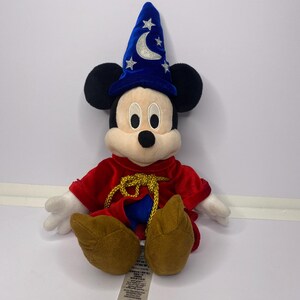 May include: A plush Mickey Mouse dressed as a sorcerer, wearing a blue hat with a crescent moon and stars. He has a red robe with gold trim and brown shoes. The toy is sitting on a white surface.