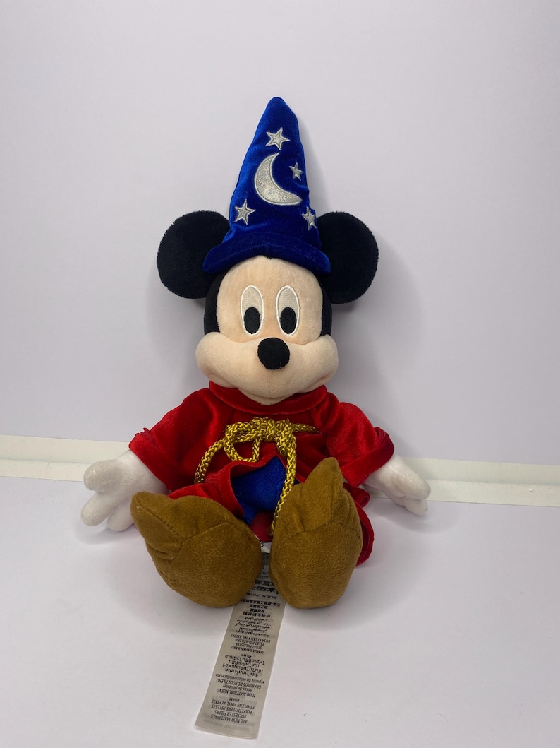 May include: A plush Mickey Mouse dressed as a sorcerer, wearing a blue hat with a crescent moon and stars. He has a red robe with gold trim and brown shoes. The toy is sitting on a white surface.
