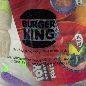May include: A white, purple, and green plush toy in a clear plastic bag. The bag has a Burger King logo and the text "This bag is not a toy. Please discard. Manufactured for Burger King Corporation. &copy; DISNEY &copy; PIXAR Made in China." The bag also has images of characters from the movie Toy Story, including Woody, Rex, and Hamm.