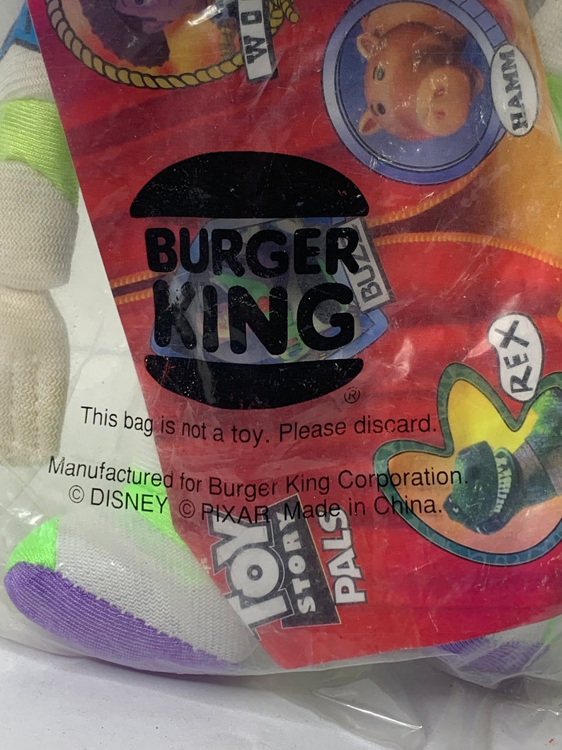 May include: A white, purple, and green plush toy in a clear plastic bag. The bag has a Burger King logo and the text "This bag is not a toy. Please discard. Manufactured for Burger King Corporation. &copy; DISNEY &copy; PIXAR Made in China." The bag also has images of characters from the movie Toy Story, including Woody, Rex, and Hamm.