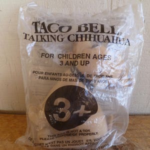 Puede incluir: Una bolsa de plástico con un juguete Taco Bell Talking Chihuahua en su interior. La bolsa es transparente y tiene el texto "Taco Bell Talking Chihuahua" y "Para niños de 3 años en adelante".