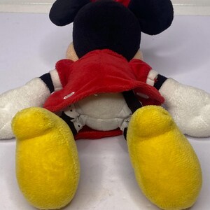 Pu&ograve; includere: Una bambola di peluche di Minnie Mouse, vista da dietro, con un vestito rosso, orecchie nere e un fiocco rosso. La bambola ha guanti bianchi e grandi piedi gialli. La bambola &egrave; sdraiata su una superficie bianca.