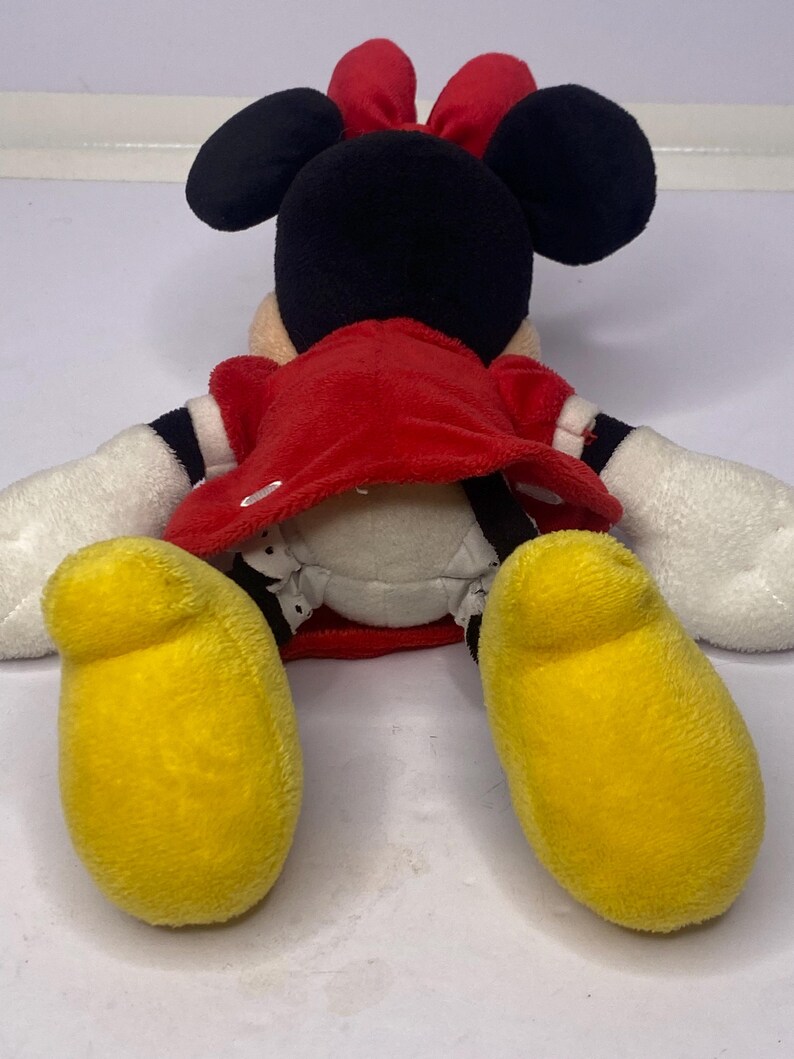 Pu&ograve; includere: Una bambola di peluche di Minnie Mouse, vista da dietro, con un vestito rosso, orecchie nere e un fiocco rosso. La bambola ha guanti bianchi e grandi piedi gialli. La bambola &egrave; sdraiata su una superficie bianca.