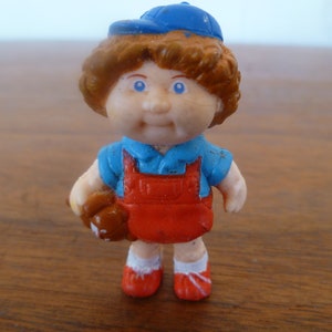 Può includere: Piccola statuetta di plastica di un bambino con berretto da baseball blu, camicia blu e tuta rossa. La statuetta tiene un guanto da baseball marrone e ha scarpe rosse. La figura ha capelli castani e occhi blu.