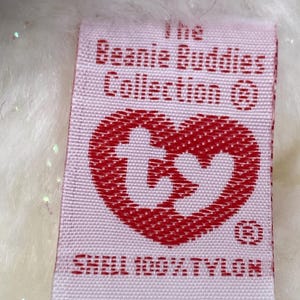 K&ouml;nnte beinhalten: Ein rechteckiges Etikett mit dem Text "The Beanie Buddies Collection" und einem roten herzf&ouml;rmigen Logo. Das Etikett lautet auch "SHELL 100% TYLON". Das Etikett ist wei&szlig; mit rotem Text und einem roten Herzen.
