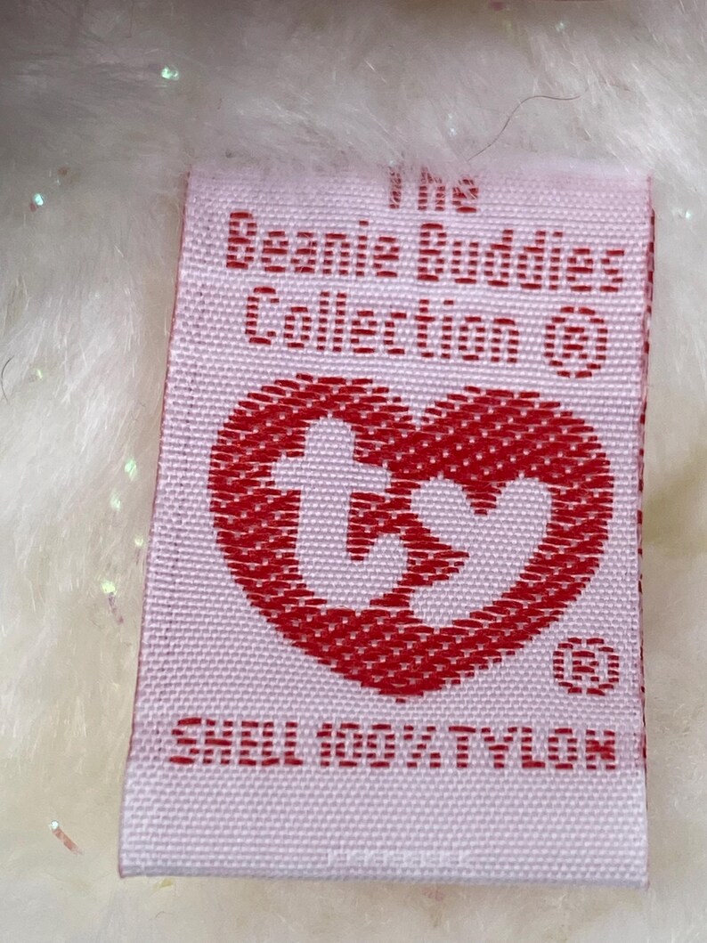 K&ouml;nnte beinhalten: Ein rechteckiges Etikett mit dem Text "The Beanie Buddies Collection" und einem roten herzf&ouml;rmigen Logo. Das Etikett lautet auch "SHELL 100% TYLON". Das Etikett ist wei&szlig; mit rotem Text und einem roten Herzen.