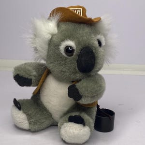 May include: A plush koala bear wearing a brown cowboy hat with the word "OUT" on it. The koala has gray fur, white accents, and black paws. It has a small black bucket attached to its back.