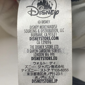 Pu&ograve; includere: Etichetta bianca con testo nero e logo Disney. L'etichetta elenca le informazioni sull'approvvigionamento e la distribuzione di prodotti Disney, inclusi i negozi Disney a Burbank, California, Londra, Inghilterra e Tokyo, Giappone.