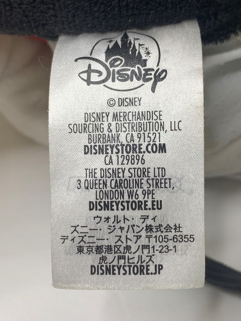 Pu&ograve; includere: Etichetta bianca con testo nero e logo Disney. L'etichetta elenca le informazioni sull'approvvigionamento e la distribuzione di prodotti Disney, inclusi i negozi Disney a Burbank, California, Londra, Inghilterra e Tokyo, Giappone.