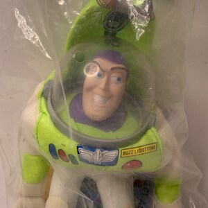 May include: A green and white plush toy of Buzz Lightyear from the Toy Story franchise. The toy is in its original packaging and has a purple face with a big smile. The packaging has the text "Buzz Lightyear" on it.