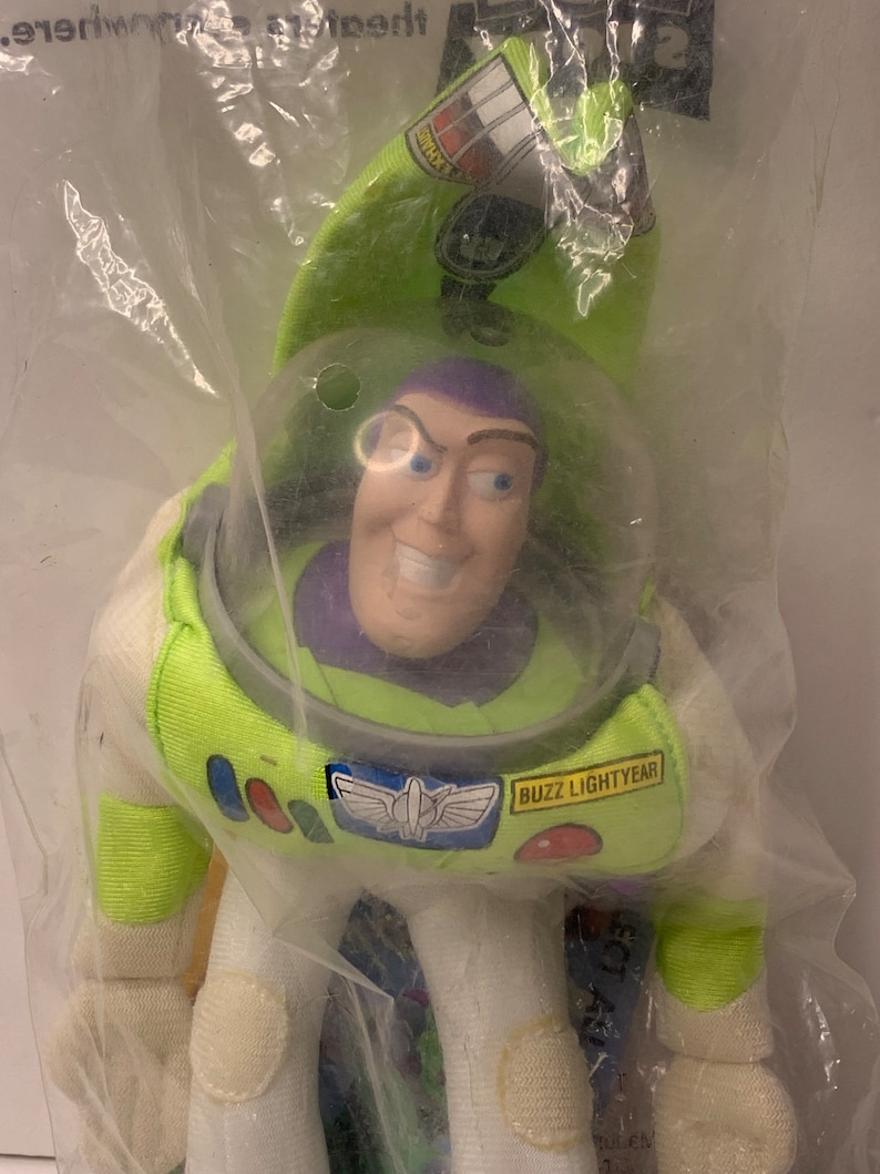 May include: A green and white plush toy of Buzz Lightyear from the Toy Story franchise. The toy is in its original packaging and has a purple face with a big smile. The packaging has the text "Buzz Lightyear" on it.