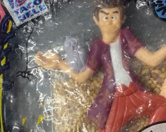Peut inclure: Un jouet Taco Bell représentant un personnage de dessin animé avec une chemise violette et un pantalon rouge. Le personnage tient un petit hibou violet. Le jouet est emballé dans un sac en plastique transparent avec un fond rose et jaune. Le sac porte le logo Taco Bell et le texte "CHOKING HAZARD" et "AVERT RISQUE IMPORTANT" imprimé dessus.